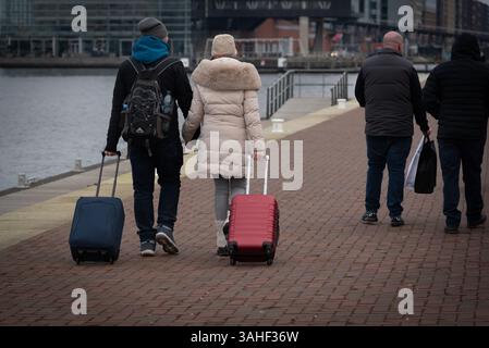 Ein Paar schlendert Hand in Hand entlang eines Uferweges und zieht ihr Gepäck, während andere an einem kühlen Tag in die entgegengesetzte Richtung gehen. Stockfoto