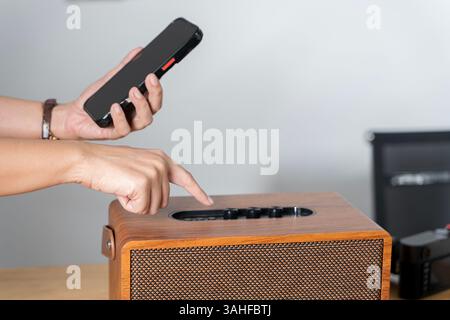 Frau, die einen intelligenten Lautsprecher mit Smartphone verwendet. Frau, die Musik auf ihrem Handy auswählt, um sie über ihren intelligenten Lautsprecher zu hören. Stockfoto