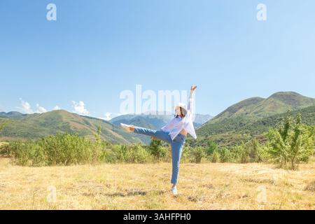 Eine fröhliche junge Frau tanzt frei auf einer goldenen Wiese, umgeben von Bäumen, mit Bergen im Hintergrund. Konzepte von Freiheit, Glück, Natur, Stockfoto