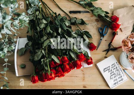Rote Rosen, Blumenwerkzeuge und ein offener Notizblock mit Kundennamen auf einer Holzarbeitsfläche. Tägliche Bestellungen, Blumengeschäft, Einzelhandel Stockfoto