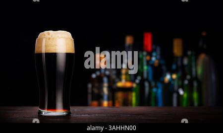Ein reichhaltiges, dunkles, kräftiges Bier, das in einem Glas auf einem rustikalen Holztisch serviert wird, mit einem warmen, verschwommenen Bokeh aus Alkoholflaschen im Hintergrund Stockfoto