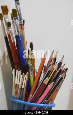 Eine Vielzahl von Kunstgegenständen ist in einem Metallbecher angeordnet und zeigt leuchtende Buntstifte und Pinsel vor einer weißen Wand. Stockfoto