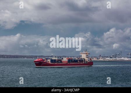 Rotes Frachtschiff, das auf einer ruhigen See unter bewölktem Himmel mit einem Hafen im Hintergrund segelt. Dänemark Stockfoto