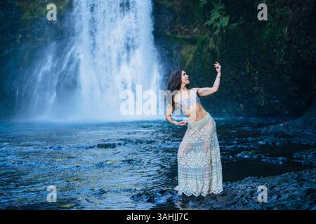 Frau in einem Spitzenrock tanzt anmutig vor einem Wasserfall in einer ruhigen Waldumgebung. Silver Falls, Oregon, USA Stockfoto