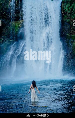 Frau in einem weißen Kleid, die in einem Pool unter einem großen, kaskadierenden Wasserfall steht. Silver Falls, Oregon, USA Stockfoto