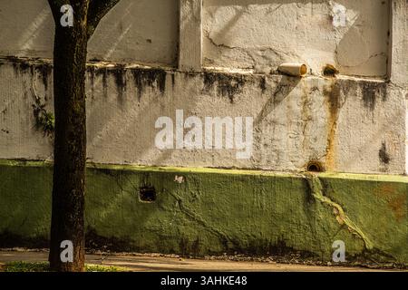 Verwitterte Stadtmauer mit abblätternder Farbe, Schimmelflecken und Abflussrohren bei Nachmittagssonne Stockfoto