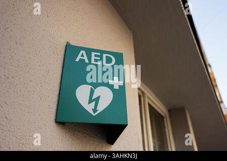 Grünes Schild mit AED-Defibrillator an der Gebäudewand, das den Standort medizinischer Notfallgeräte anzeigt. Stockfoto