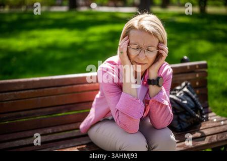 Die nachdenkliche reife Frau sitzt an sonnigen Tagen auf einer Bank im Park. Stockfoto