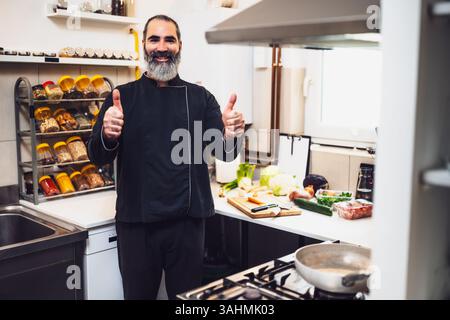 Porträt eines professionellen Küchenchefs, der in der Küche des Restaurants arbeitet. Er ist bereit, eine Mahlzeit zuzubereiten. Koche in der Küche und zeige die Daumen hoch. Stockfoto