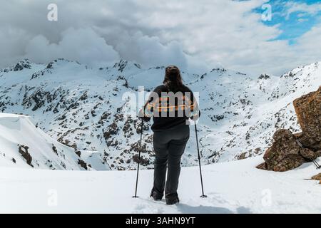 Eine Frau steht selbstbewusst auf einem schneebedeckten Berghang, hält Wanderstöcke und blickt auf die schneebedeckte Sierra de Gredos in Spanien. Gekleidet Stockfoto