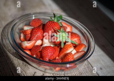 Frische Erdbeeren (Fragaria x anananassa) in einer Glasschale auf rustikalem Holz, geschnitten und ganz, ideal für gesunde Ernährung, Sommerrezepte und Lifestyle foo Stockfoto