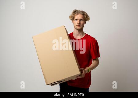 Junger Liefermann, der einen großen Karton auf weißem Hintergrund hält. Stockfoto