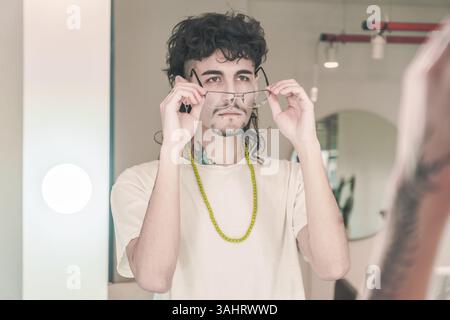 Ein stilvoller junger Mann mit lockigen Haaren und einem Spitzbart probiert eine neue Brille an, während er in den Spiegel eines optischen Ladens blickt und sie mit einer neuen Brille anpasst Stockfoto