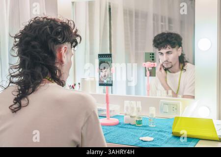 Junger Mann mit einer trendigen Meeräsche, der sich selbst filmt, während er Make-up vor einem Spiegel aufträgt, mit einem Smartphone, das auf einem Stativ in einer vibra montiert ist Stockfoto