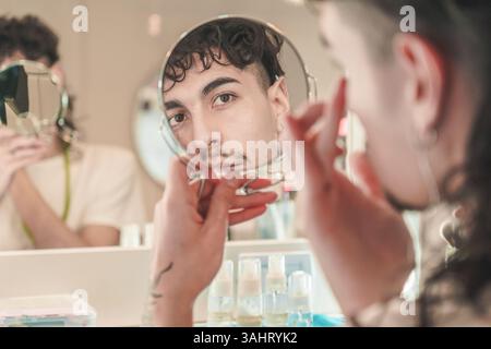 Junger Mann, der sein Gesicht im Spiegel reflektiert, während er Cremes für seine tägliche Hautpflege im Badezimmer aufträgt, während eine andere Person aus ihm raus ist Stockfoto