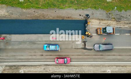 Blick aus der Vogelperspektive auf Straßenpflaster mit Verkehr in der Nähe. Luftaufnahme von Arbeitern, die mit Maschinen eine Straße mit frischem Asphalt pflastern, während Autos nebenan fahren Stockfoto