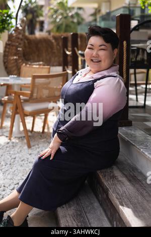 Eine junge asiatische Frau in Übergröße sitzt selbstbewusst vor einem Café und trägt ein stilvolles Outfit. Sie legt ihr Kinn auf die Hand, während sie zur Seite blickt. Stockfoto