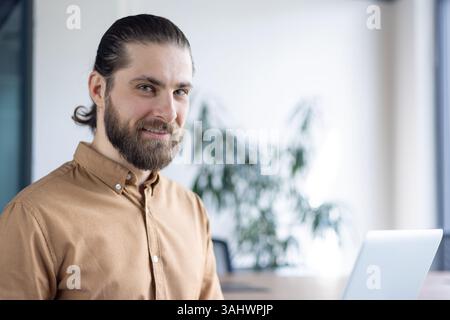 Ein lächelnder Geschäftsmann mit Bart schaut in einem Büro zur Kamera. Er trägt ein braunes Hemd und sitzt neben einem Laptop auf einem Schreibtisch. Stockfoto