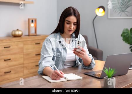 Schöne Frau mit Planer, die Notizen macht und Smartphone am Holztisch im Haus benutzt Stockfoto