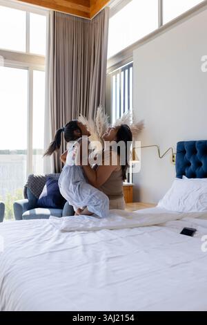 Glückliche Mutter und Tochter genießen einen verspielten Moment drinnen im Schlafzimmer mit Smartphone und Bettwäsche. Familie, Bindung, Glück, Verspieltheit, lässig, gemütlich Stockfoto