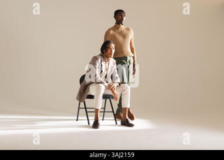 afroamerikaner sitzt auf einem Stuhl in einem minimalistischen Studio mit asiatischem Mann. studio, minimalistisch, Beleuchtung, Selbstvertrauen, Teamwork, Freundschaft, fa Stockfoto