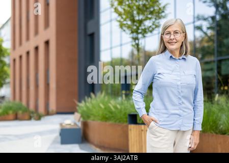 Eine elegante Seniorin steht selbstbewusst draußen, in der Nähe eines modernen Gebäudes mit grünen Akzenten, und lächelt sanft. Ihr Outfit ist schick und professionell. Stockfoto
