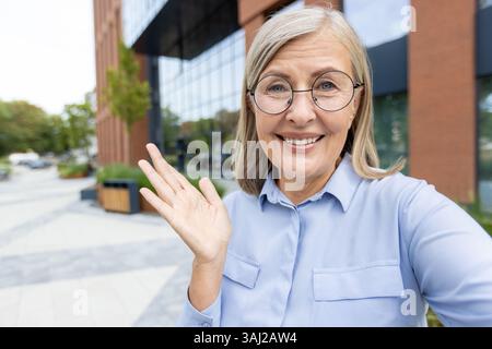 Eine ältere Frau, die eine runde Brille trägt, lächelt und Gesten vor der Kamera draußen in der Nähe eines modernen Gebäudes mit Glasfenstern. Stockfoto