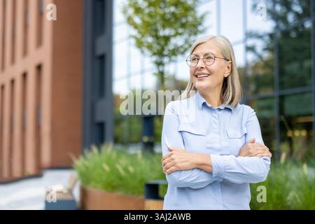 Eine lächelnde, reife Geschäftsfrau steht draußen mit überkreuzten Armen in der Nähe eines Bürogebäudes. Sie blickt auf etwas mit Optimismus. Stockfoto