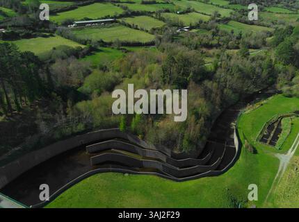 Ein hochwinkliger Luftschuss fängt ein Betonwehr ein, dessen Wasser in mehreren Ebenen in einen Kanal fließt, umgeben von üppigen grünen Feldern und Bäumen Stockfoto