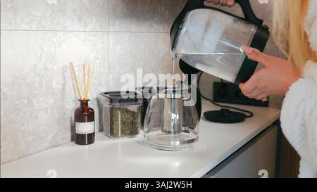 Frau gießt heißes Wasser in eine Teekanne aus Glas. In einer modernen Kücheneinrichtung wird eine Frau gesehen, die heißes Wasser in eine transparente Teekanne aus Glas gießt. Stockfoto