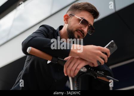 Ein junger Hipster-Mann mit einem Elektroroller, der vor einem modernen Geschäftsgebäude auf der Stadtstraße steht und ein Mobiltelefon benutzt. Stockfoto