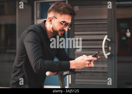 Ein junger, stilvoller Mann mit elektrischem Roller, der vor einem modernen Geschäftsgebäude in der Stadtstraße steht und ein Telefon benutzt. Stockfoto