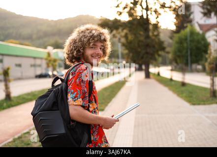 Ein glücklicher Student auf einem Universitätscampus draußen, der die Straße entlang läuft, auf die Kamera zurückblickt und lächelt. Stockfoto