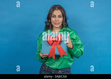 Eine junge Frau zeigt eine blaue Geschenkbox mit roter Schleife und feiert einen besonderen Anlass Stockfoto