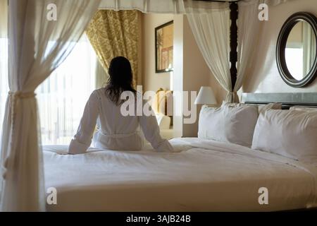 Eine Person in einem Bademantel sitzt auf einem sauber gemachten Bett und blickt aus einem großen Fenster. Sanftes Sonnenlicht erfüllt den Raum und schafft eine friedliche und ruhige Atmosphäre Stockfoto