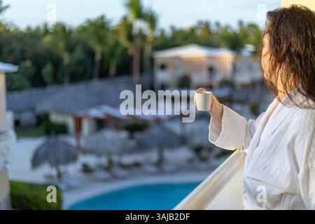 Eine Frau im Bademantel genießt eine Tasse Kaffee auf dem Balkon. Die Aussicht umfasst einen Swimmingpool und üppiges Grün in einem tropischen Resort am Morgen Stockfoto