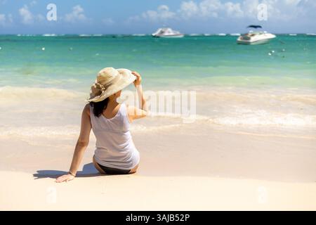 Eine Frau sitzt am Sandstrand und blickt auf das ruhige Meer. Sie trägt einen Strohhut, wenn die Sonne hell scheint, mit Booten am Horizont aga Stockfoto
