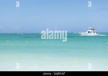 Ein weißes Boot gleitet sanft durch das türkisfarbene Wasser in der Nähe eines Sandstrandes unter klarem Himmel. An einem sonnigen Tag schlängeln sich die Wellen sanft am Ufer. Stockfoto