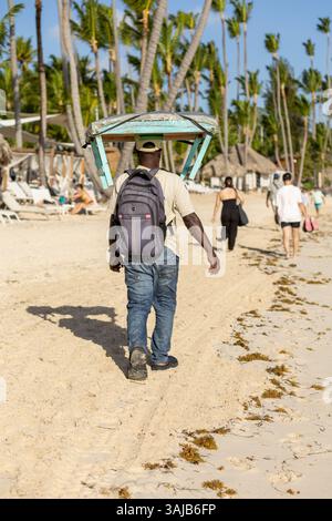 Punta Cana, Dominikanische Republik, 25. märz 2025: Ein Mann läuft an einem Sandstrand entlang und balanciert ein Holzboot auf seinem Kopf. Palmen schwanken im Hinterland Stockfoto