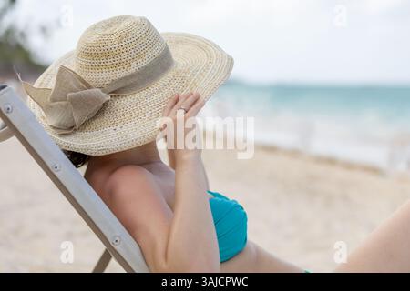 Eine Frau genießt einen ruhigen Moment am Strand, lehnt sich auf einem Stuhl, trägt einen großen Sonnenhut und einen Bikini. Die Wellen des Ozeans schlängeln sanft an der Ufer Stockfoto