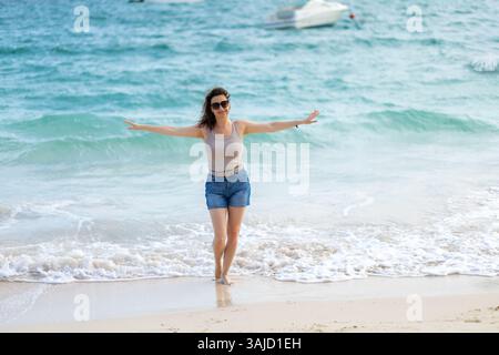 Eine Frau steht im flachen Wasser und lächelt, während sie ihre Arme ausstreckt. Mit einem Tanktop und Jeansshorts genießt sie die warme Sonne und das sanfte Meer Stockfoto
