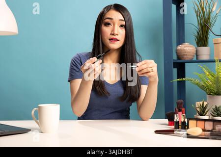Junge Make-up-Künstlerin hält eine Mascara und filmt ein Tutorial für ihre Online-Zuschauer. Asiatische Vloggerin sitzt am Schreibtisch und nimmt Livestream auf, um ihre Schönheitsroutine im Studio mit blauem Hintergrund zu präsentieren. Stockfoto