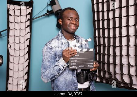 Ein männlicher afroamerikanischer Regisseur posiert selbstbewusst in einem Studio mit professioneller Beleuchtung, hält eine Trennwand und eine DSLR. Schwarzer Fotograf lächelt in die Kamera mit einem Filmschiefer in der Hand. Stockfoto