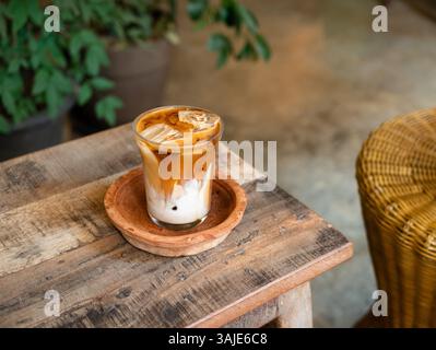 Eiskaffee serviert in einem Glas mit Milch und Espresso auf einem rustikalen Holztisch in einem gemütlichen Café mit natürlichem Licht und grünen Pflanzen Stockfoto