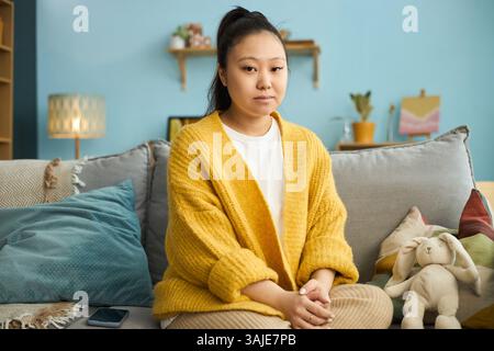 Porträt einer asiatischen Frau, die auf dem Sofa sitzt, in gelbem Cardigan, während sie die Kamera ansieht, mit gemütlichem Ambiente, einschließlich Kissen, Lampe und Pflanze Stockfoto