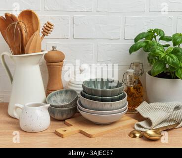 Verschiedene Küchenutensilien, darunter Schüsseln, Holzlöffel, Pfeffermühle, Ölflaschen, und eine Basilikumpflanze, die ordentlich auf einer hölzernen Arbeitsplatte angeordnet sind Stockfoto