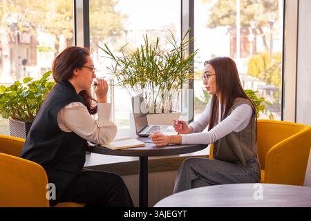 Zwei selbstbewusste Brünetten in einem Café, die neue Funktionen für ihr digitales Start-up, das Konzept der Geschäftsstrategie und Innovation finden. Mitten im Shot. Stockfoto