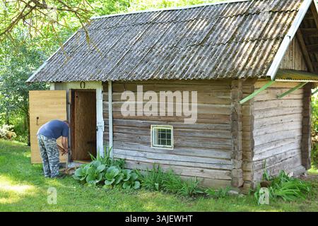 Älterer Mann, der Stämme mit Axt für altes Badehaus im ländlichen Raum Litauen spaltet. Senior-Mann, der altes Badehaus für die Benutzung vorbereitet. Kleine Hütte auf dem Lande in der EU Stockfoto