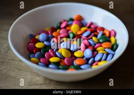 Eine volle Schüssel mit bunten Süßigkeiten aus nächster Nähe auf einem Holztisch. Schokoladenknöpfe süße Leckereien in einer weißen Schüssel. Stockfoto
