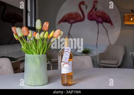 Eine gemütliche und stilvolle Inneneinrichtung mit einer grünen Vase mit orangefarbenen und weißen Tulpen, einer Flasche Rosenwein zur Begrüßung und einem weichen Hintergrund mit rosa Stockfoto